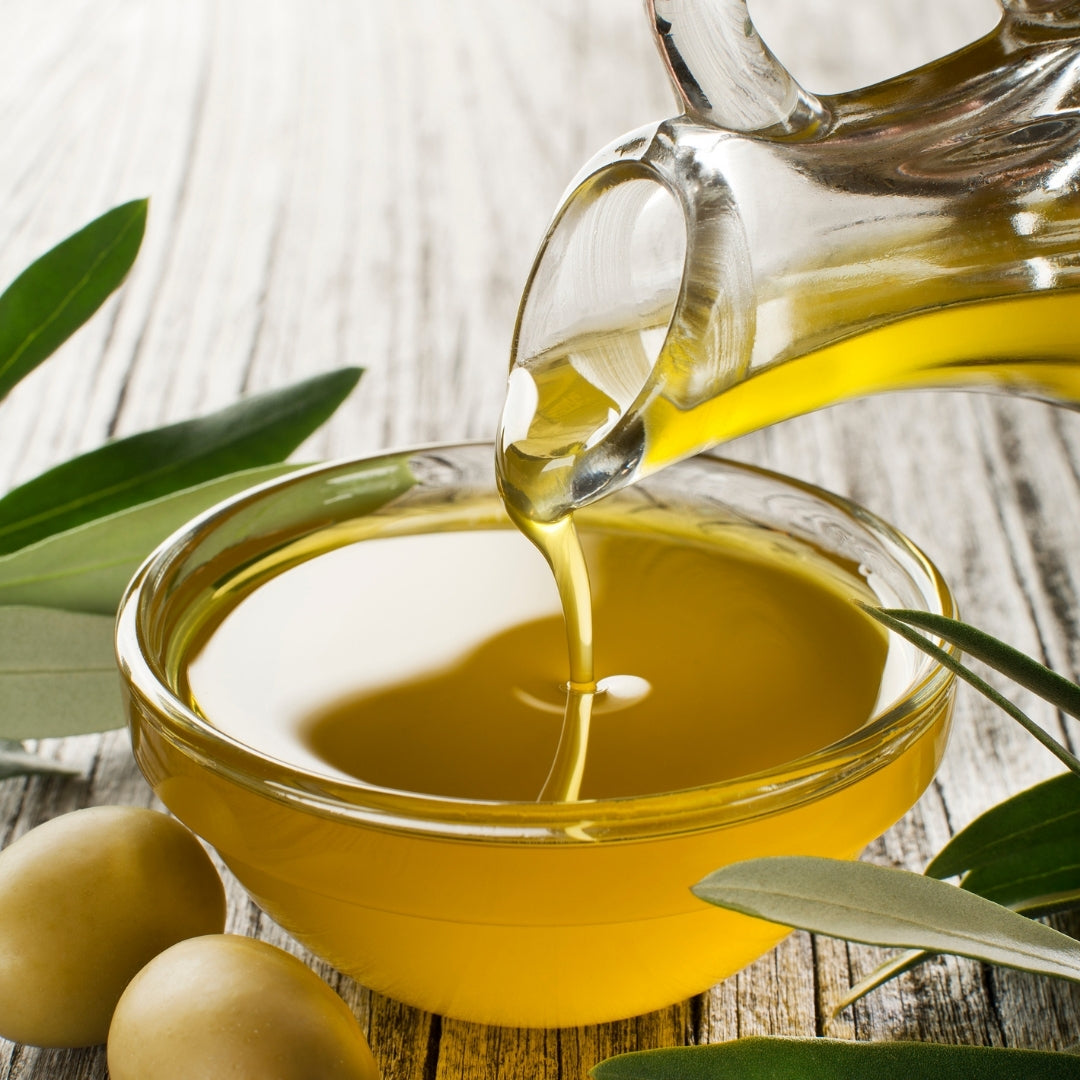 Olive oil being poured from a glass container into a bowl on a wooden surface with olive leaves.