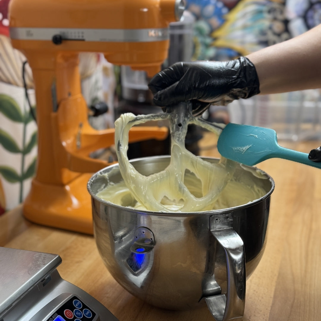 Person using a blue spatula to remove tallow moisturizer batter from a stand mixer bowl with an orange stand mixer in the background.